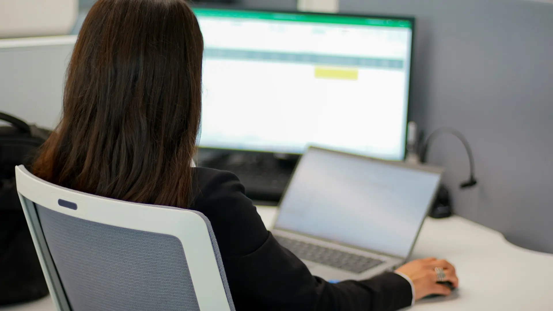 Vue de dos d'une femme travaillant devant un ordinateur de bureau et un ordinateur portable dans un espace de travail.