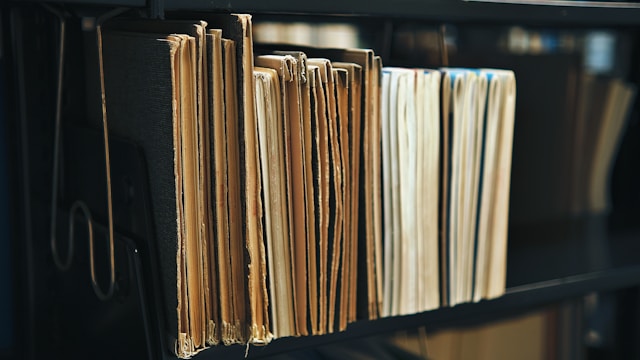 De vieux dossiers en carton et des classeurs de factures alignés sur une étagère de bureau noire, symbolisant le tri et le nettoyage d'une balance âgée ou d'un poste client laissé à l'abandon.
